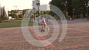 A little boy in a red checkered shirt rides a scooter in a city Park at sunset.