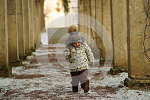 Little boy with red cheeks, in bright clothes and mittens walking up the path and looking down in snowy winter park
