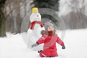 Little boy in red clothes having fun with big snowman. Child during stroll in a snowy winter park. Active winter outdoor leisure