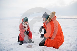 A little boy in a red jumpsuit and scarf is fishing in an ice hole with his father