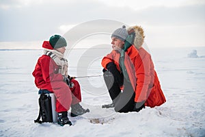 A little boy in a red jumpsuit and scarf is fishing in an ice hole with his father