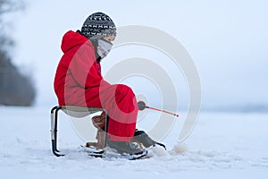 Little boy in red overalls on winter fishing. Sits on a folding chair. In the hands of a fishing rod.