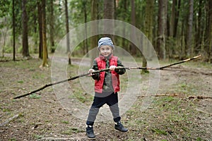 Little boy in red vest is playing with big branch and having fun in forest on early spring day. Outdoor activity for children
