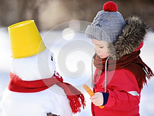 Little boy in red winter clothes having fun with snowman in snow