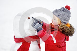 Little boy in red winter clothes having fun with snowman in snowy park