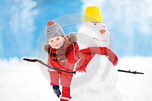 Little boy in red winter clothes having fun with snowman in snowy park