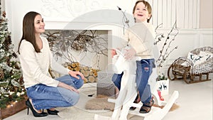 Little boy riding wooden rocking horse and talking to his mother sitting near the Christmas tree