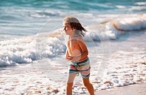 Little boy running on beach shore splashing water in blue sea. Kid walking the summer beach.