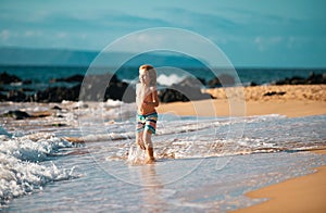 Little boy running on beach shore splashing water in blue sea. Kid walking the summer beach.