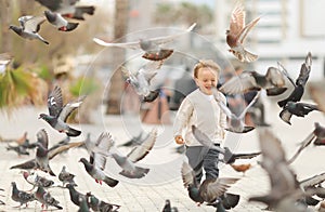 Little boy running near doves, chasing pigeons, happy child with smiling face. Kid is feeding pigeons in city park