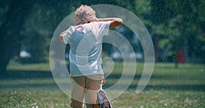 Little boy running under sprinklers in park hiding ears from water droplet alone