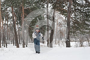 Little boy runs merrily through snow. Child plays in winter forest on large fir trees background