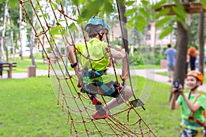 Little boy in safety equipment climbing on rope wall at adventure park. Fiend making a photo shot on smartphone. Children summer s