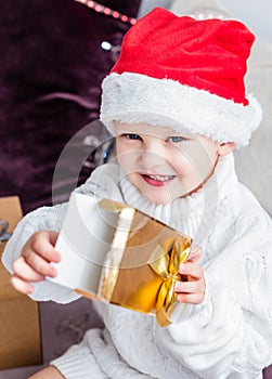 A little boy in a Santa Claus hat sits on the couch with an open gift in his hands in a golden box