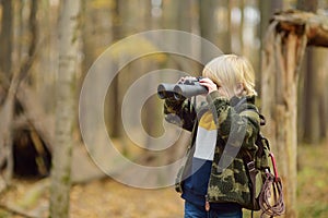 Little boy scout with binoculars during hiking in autumn forest. Child looking through a binoculars. Behind the baby is teepee hut