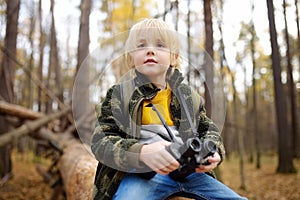Little boy scout with binoculars during hiking in autumn forest. Child is sitting on large fallen tree and looking through a