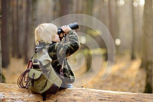 Little boy scout with binoculars during hiking in autumn forest. Child is sitting on large fallen tree and looking through a