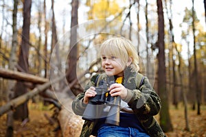 Little boy scout with binoculars during hiking in autumn forest. Child is sitting on large fallen tree and looking through a