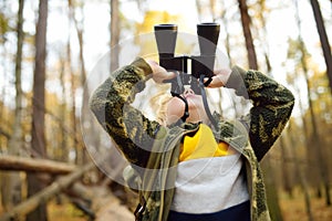 Little boy scout with binoculars during hiking in autumn forest. Child is sitting on large fallen tree and looking through a