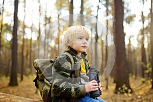 Little boy scout with binoculars during hiking in autumn forest. Child is sitting on large fallen tree and looking through a