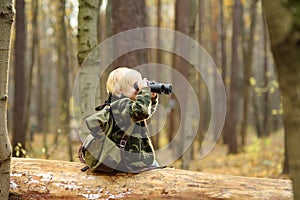 Little boy scout with binoculars during hiking in autumn forest. Child is sitting on large fallen tree and looking through a