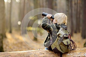 Little boy scout with binoculars during hiking in autumn forest. Child is sitting on large fallen tree and looking through a