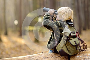 Little boy scout with binoculars during hiking in autumn forest. Child is sitting on large fallen tree and looking through a