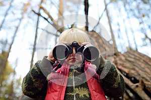 Little boy scout is looking through binoculars during hiking in autumn forest. Behind the child is teepee hut