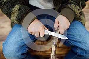 Little boy scout is sharpening a stick with the help knife in the forest