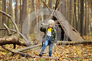 Little boy scout with spyglass during hiking in autumn forest. Child is looking through a spyglass. Behind the baby is teepee hut