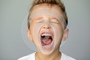 Little boy screams with closed eyes. Isolated young man on gray background opened his mouth wide