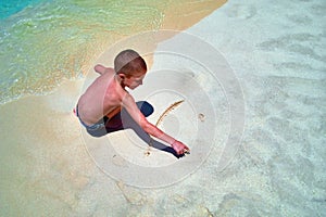 Little boy on seashore paints a smiling sun on sand. Cute child paints smiley face on sand in surf line. Top view, copy space.