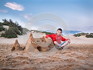 Little boy in shorts and a jacket sitting legs crossed near the sandy castle against the blue sky and the sand dunes