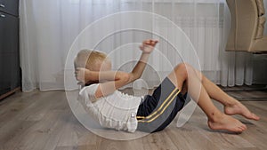 A little boy in shorts and a T-shirt shakes his abs lying on the floor at home.