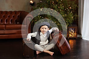 Little boy sit on wooden floor with christmas suitcase in Cozy living room. Smiling Little blond boy in white shirt with vest and