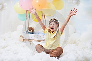 A little boy sits in a balloon basket in the clouds, pretending to travel and fly with an Aviator hat for a concept of creativity