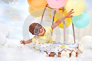 A little boy sits in a balloon basket in the clouds, pretending to travel and fly with an Aviator hat for a concept of creativity