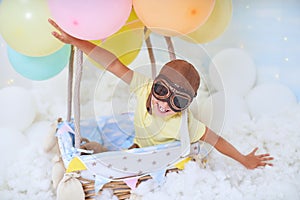 A little boy sits in a balloon basket in the clouds, pretending to travel and fly with an Aviator hat for a concept of creativity