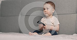 The little boy sits on the bed and watching TV. Baby watches cartoons with remote control in hands. Slow motion.