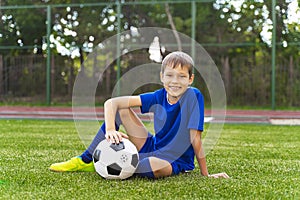 A little boy sits on a green soccer field, a soccer ball