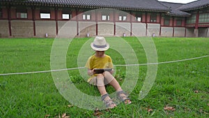 A little boy sits on a lawn in an ancient Korean palace and plays a tablet. The contradiction of modern technologies and