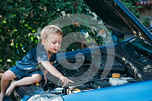 The little boy sits on the open hood of the car and examines the motor. A child studies the car and plays in the car