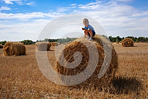 Little boy sits on a round haystack. Field with round bales after harvest under blue sky