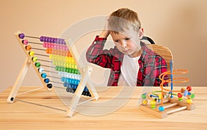 A little boy sits at the table and plays with wooden educational toys, scratches his head and thinks about the task.