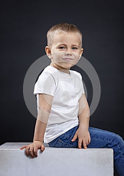 A little boy sits on a white cube. Smiling child in jeans and a white T-shirt. Black background. Vertical