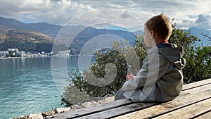 Little boy sits on wooden bench looking at Adriatic sea bay