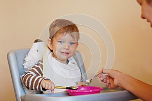 Little boy sitting at a chair for feeding and smiles. He holds a spoon in his hand and tries to eat himself from the plate