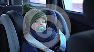 A little boy is sitting in a child seat in a car. A baby travels in a car in winter. Boy in jacket and hat.