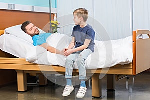 Little boy sitting on hospital bed and looking at sick father