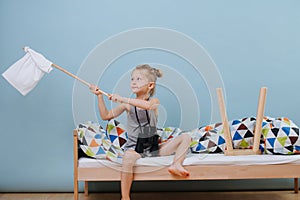 Little boy is sitting on unmade bed, waving white flag over blue walls